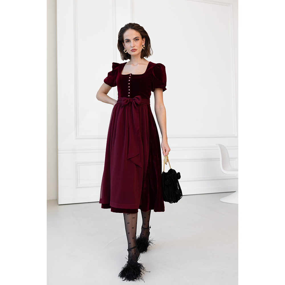 Auffälliges rotes Samt-Dirndl mit modernem Mieder, geradem Ausschnitt und Knopfleiste. Die abnehmbare Schürze ist mit einer Schleife gebunden, das Dirndl endet knapp über dem Knie.