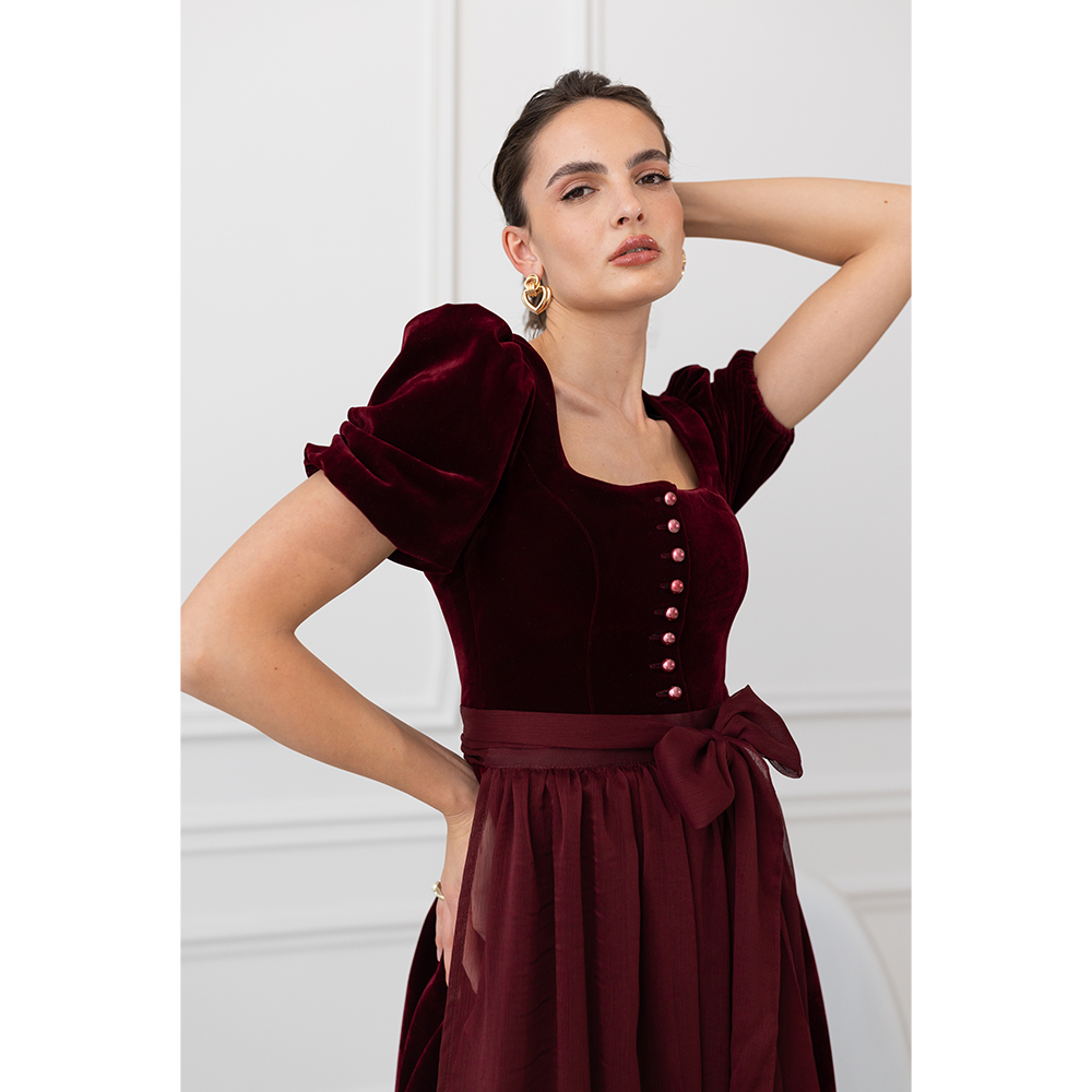 Auffälliges rotes Samt-Dirndl mit modernem Mieder, geradem Ausschnitt und Knopfleiste. Die abnehmbare Schürze ist mit einer Schleife gebunden, das Dirndl endet knapp über dem Knie.