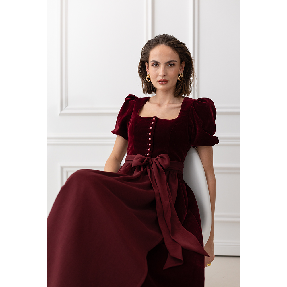 Auffälliges rotes Samt-Dirndl mit modernem Mieder, geradem Ausschnitt und Knopfleiste. Die abnehmbare Schürze ist mit einer Schleife gebunden, das Dirndl endet knapp über dem Knie.