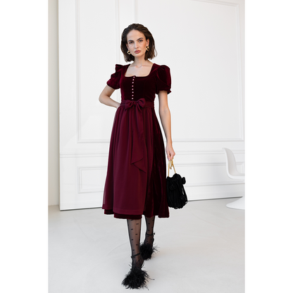 Auffälliges rotes Samt-Dirndl mit modernem Mieder, geradem Ausschnitt und Knopfleiste. Die abnehmbare Schürze ist mit einer Schleife gebunden, das Dirndl endet knapp über dem Knie.