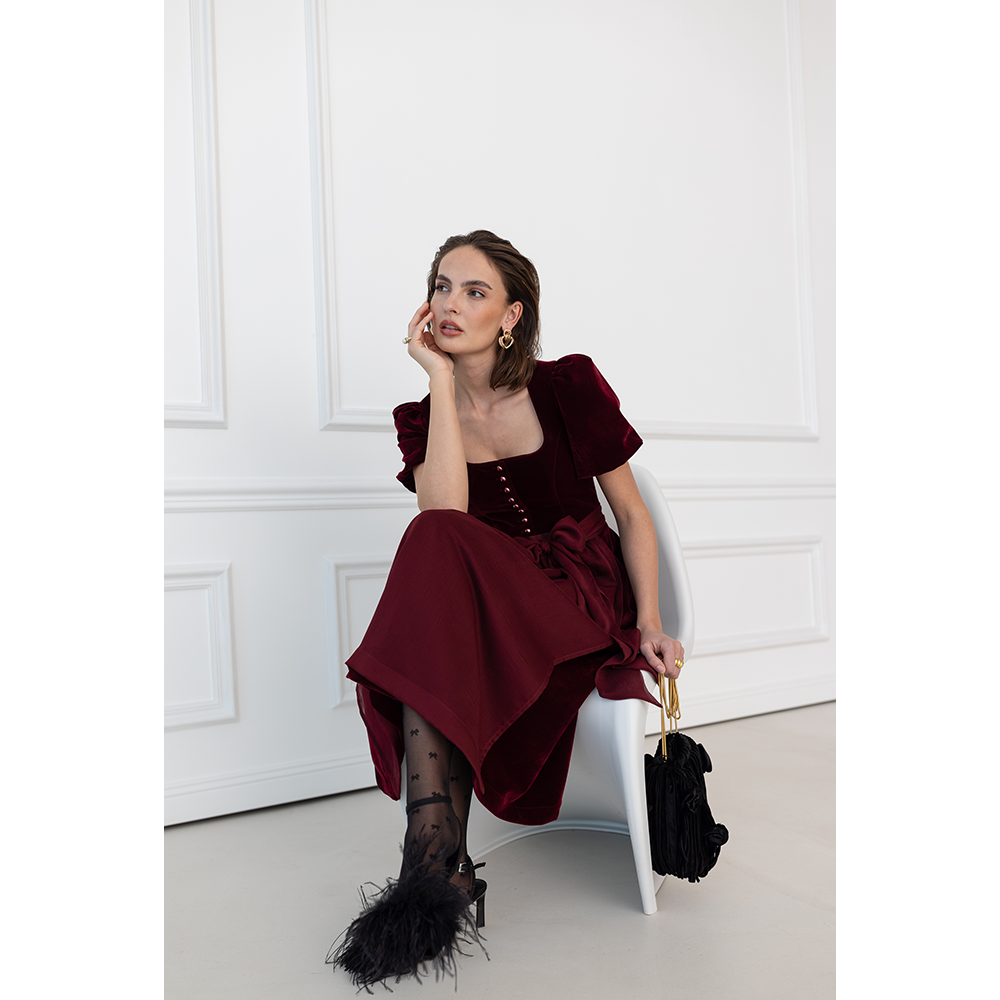 Auffälliges rotes Samt-Dirndl mit modernem Mieder, geradem Ausschnitt und Knopfleiste. Die abnehmbare Schürze ist mit einer Schleife gebunden, das Dirndl endet knapp über dem Knie.